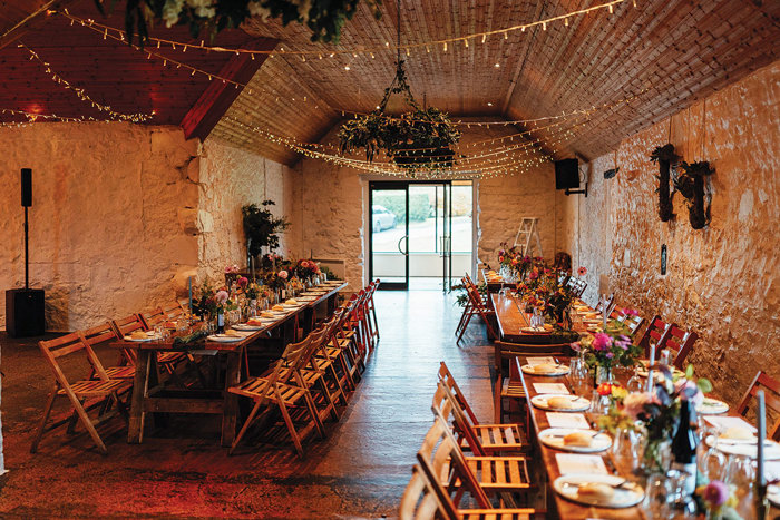 several long wooden tables all with foldable wooden chairs and table settings, are decorated with flowers and candlesticks in a stone room space draped in fairylights