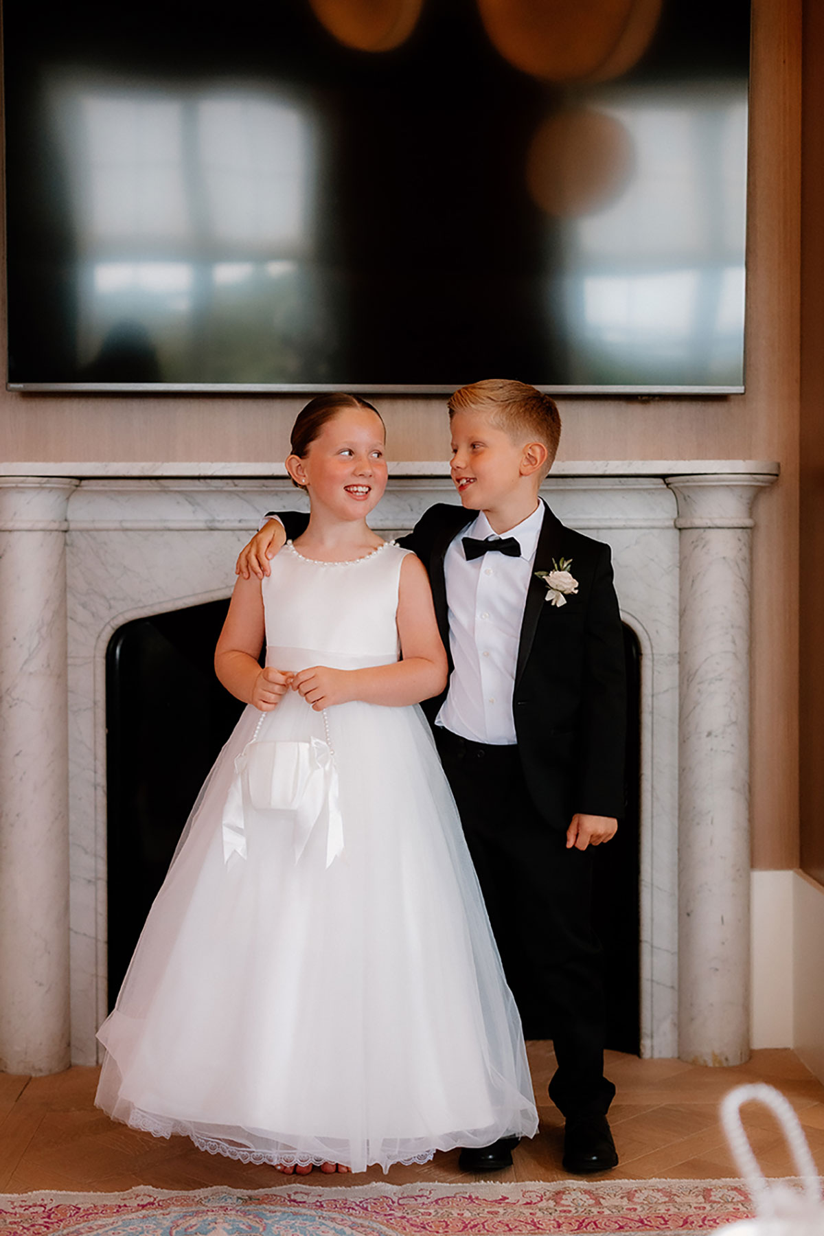Flower girl and page boy smiling together in formal outfits in front of a marble fireplace at Mar Hall.