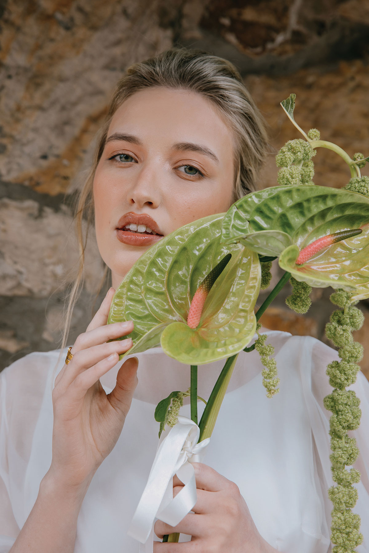 A close-up of a bride in a white gown holding a bouquet of large green anthurium flowers in front of a rustic stone wall.