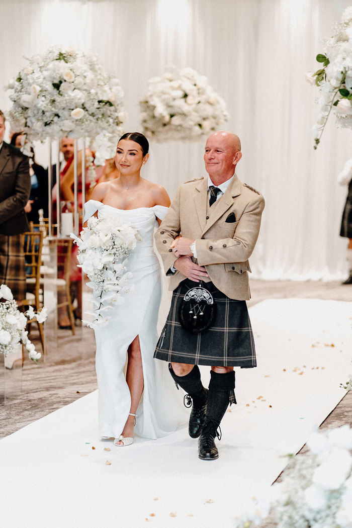 bride in strapless wedding dress with off the shoulder straps and long veil holds trailing bouquet while walking down aisle with man father figure in brown jacket and dark coloured kilt