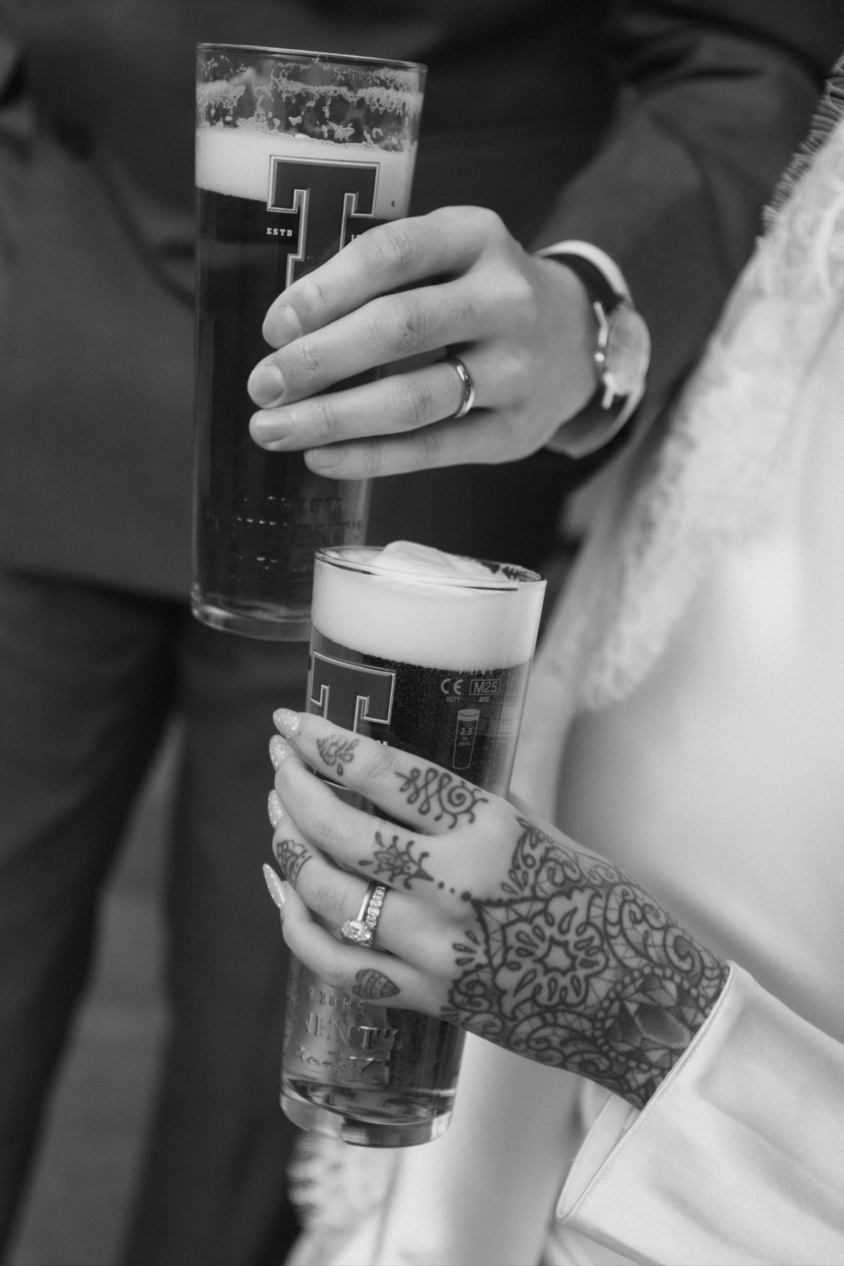 Black-and-white close-up of the newlyweds holding pints, showing their wedding rings and the bride’s hand tattoos.
