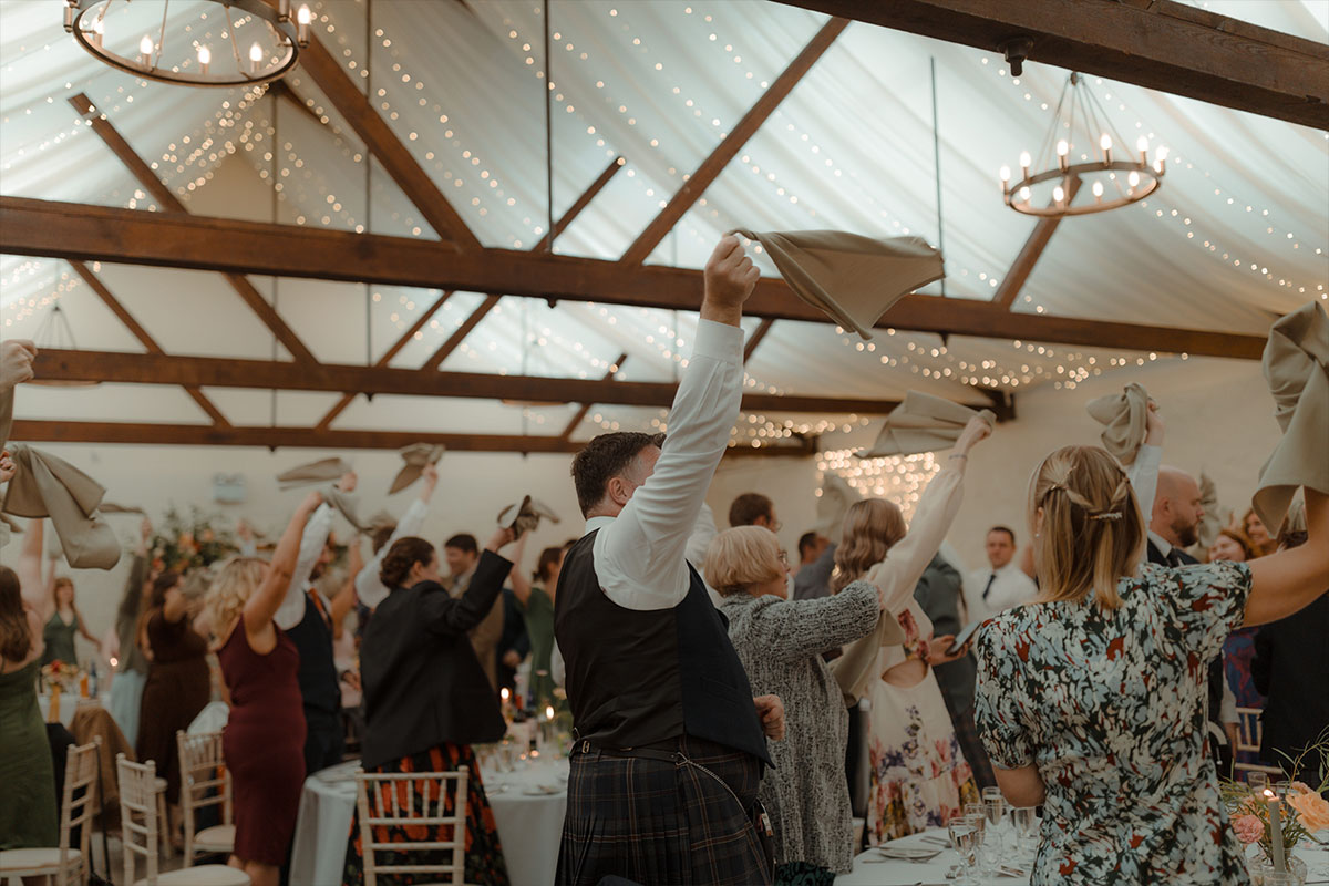 wedding guests swinging their napkins above their head while standing up inside a barn that has white fabric and fairylights draped from the roof