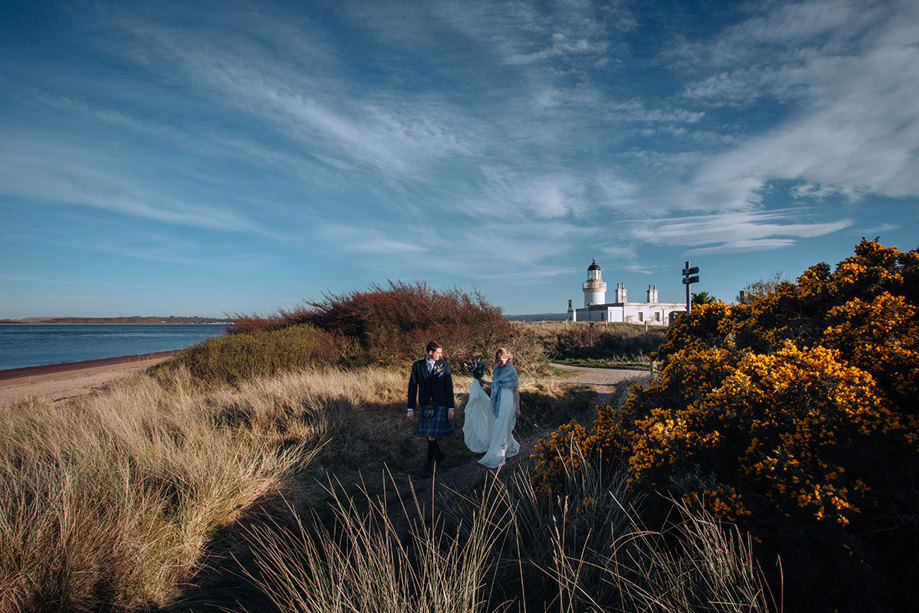 Bride and groom walking on a beach with lighthouse in background