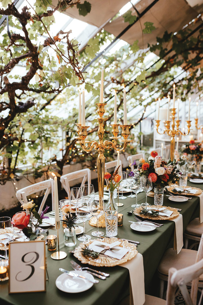 greenary filled dining space has long table with green tablecloth, lots of colourful floral arrangements, gold charger plates and tall shiny golden candelabras