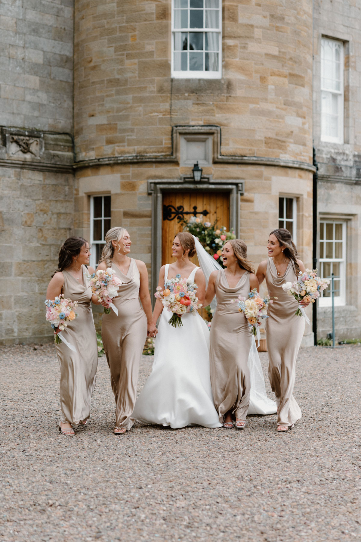 a bride and bridesmaids walking towards the camera smiling and laughing with a castle behind them