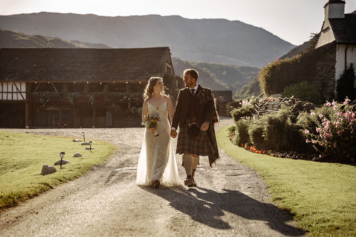 Bride and groom walk hand in hand along a country path at sunset, with barns and hills in the background. The bride wears a lace wedding dress and carries a wildflower bouquet, while the groom wears a brown tweed jacket with tartan kilt and fly plaid