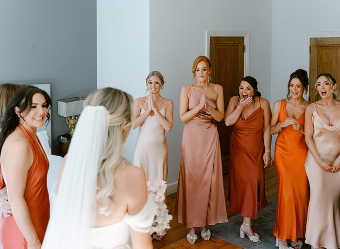 a bride revealing her dress to 6 surprised-looking bridesmaids wearing orange, terracotta and oyster-coloured dresses