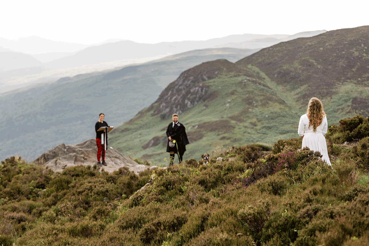 Bride approaches her groom and celebrant across a rugged hillside during an intimate Scottish mountain elopement with panoramic Highland views
