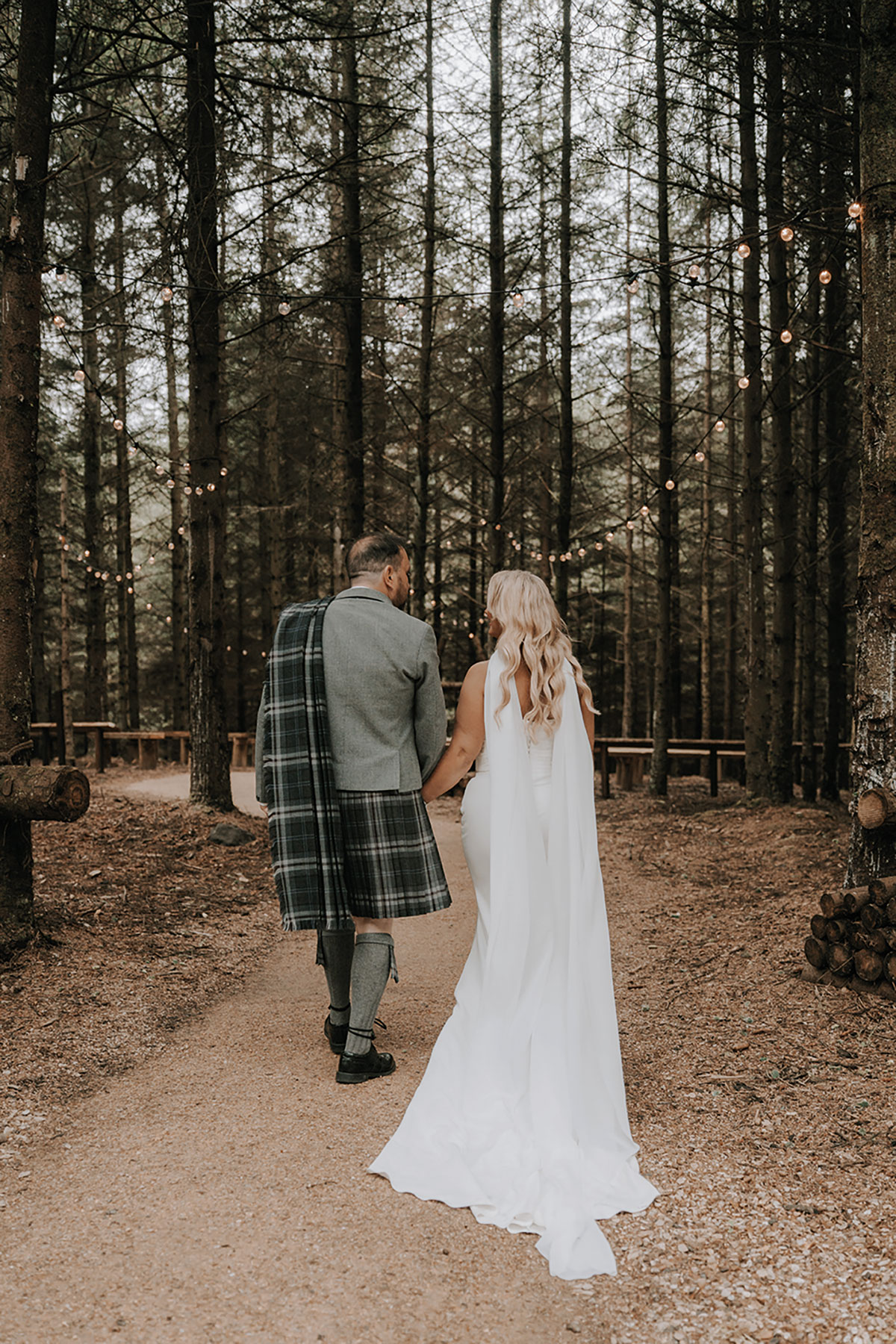 Bride and groom walking hand in hand along a forest path lined with string lights.