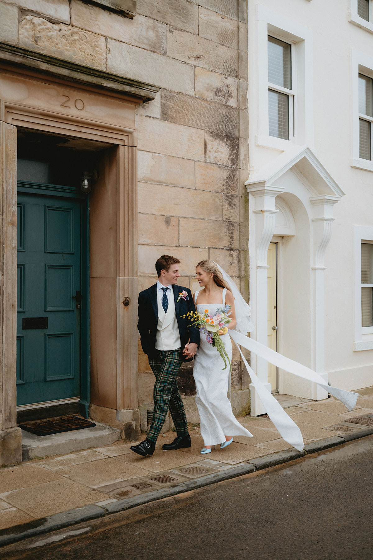 Newlywed couple walking along a quiet Scottish street as the bride’s long veil trails behind them.