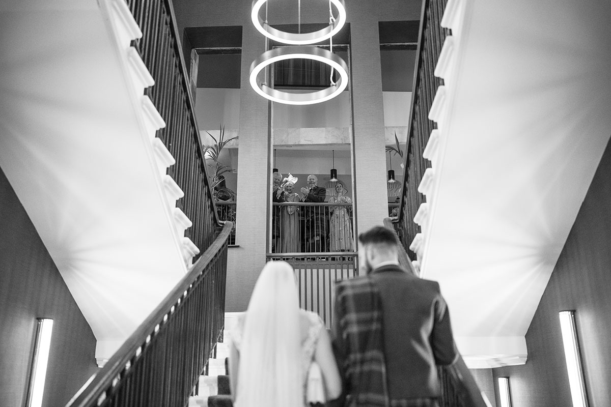 Bride and groom walk up the staircase at Clayton Hotel Glasgow as guests watch from the balcony.