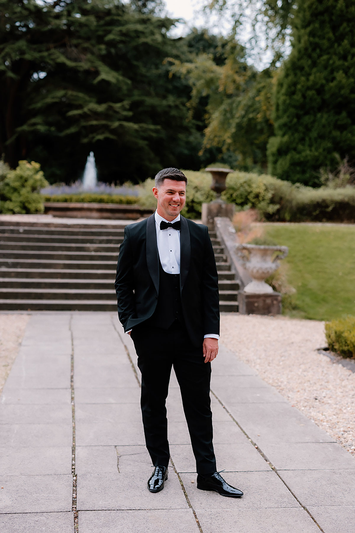 Groom standing on the garden pathway at Mar Hall, smiling with the fountain and steps behind him.