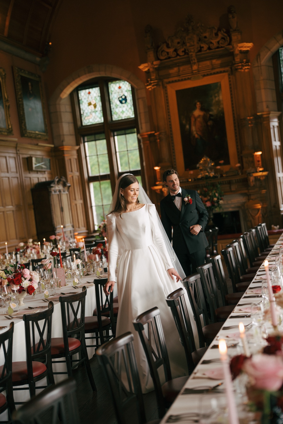 Nicole and Ryan admire their wedding reception setup at Barnbougle Castle, featuring long dining tables decorated with pink-and-red flowers and candles.