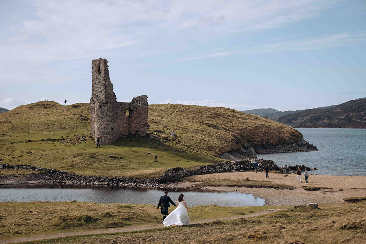 Bride and groom walk hand in hand near the ruins of Ardvreck Castle overlooking Loch Assynt after their Achnahaird Bay elopement.