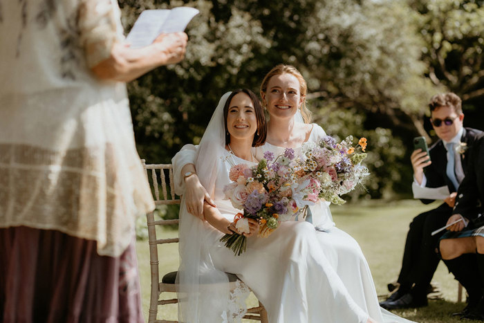 two smiling brides seated holding large pastel bouquets in the walled garden at the Byre at Inchyra