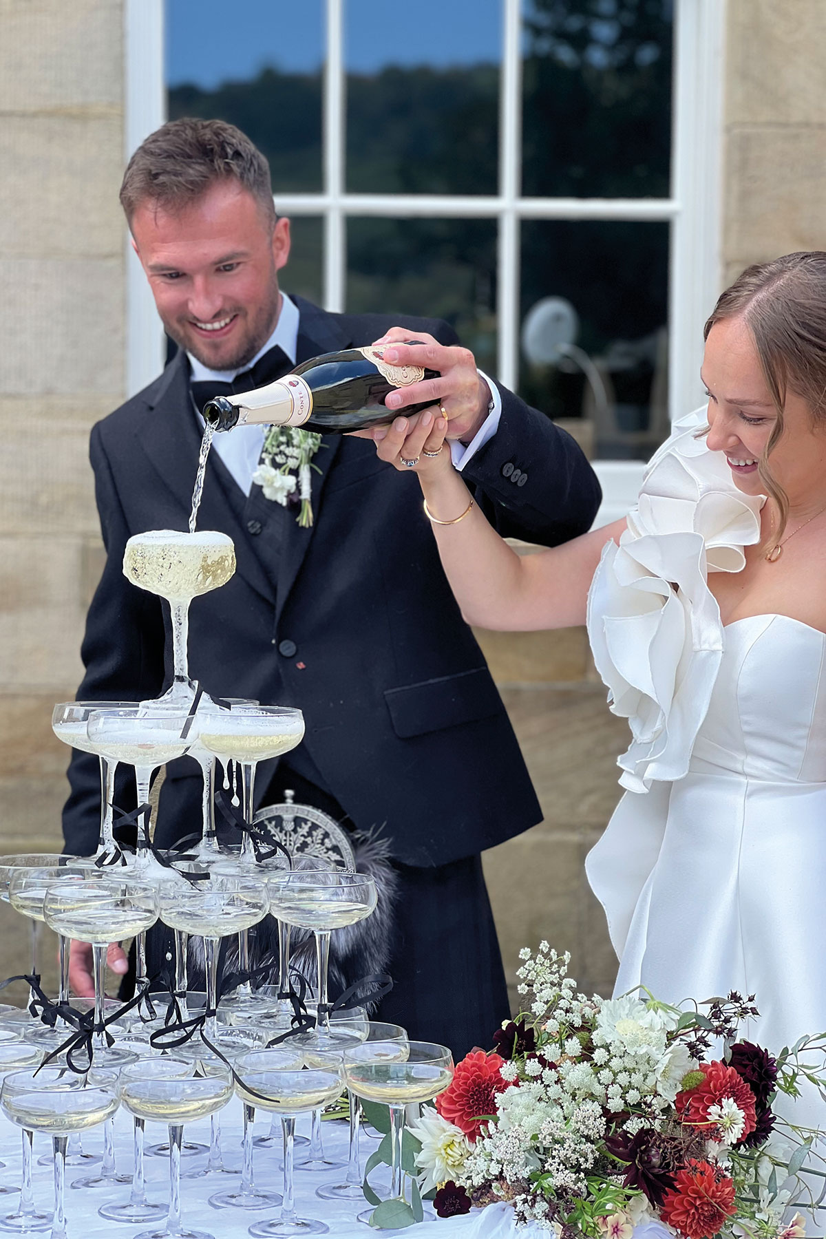 Bride and groom pouring champagne into a champagne tower surrounded by colourful wedding flowers outdoors