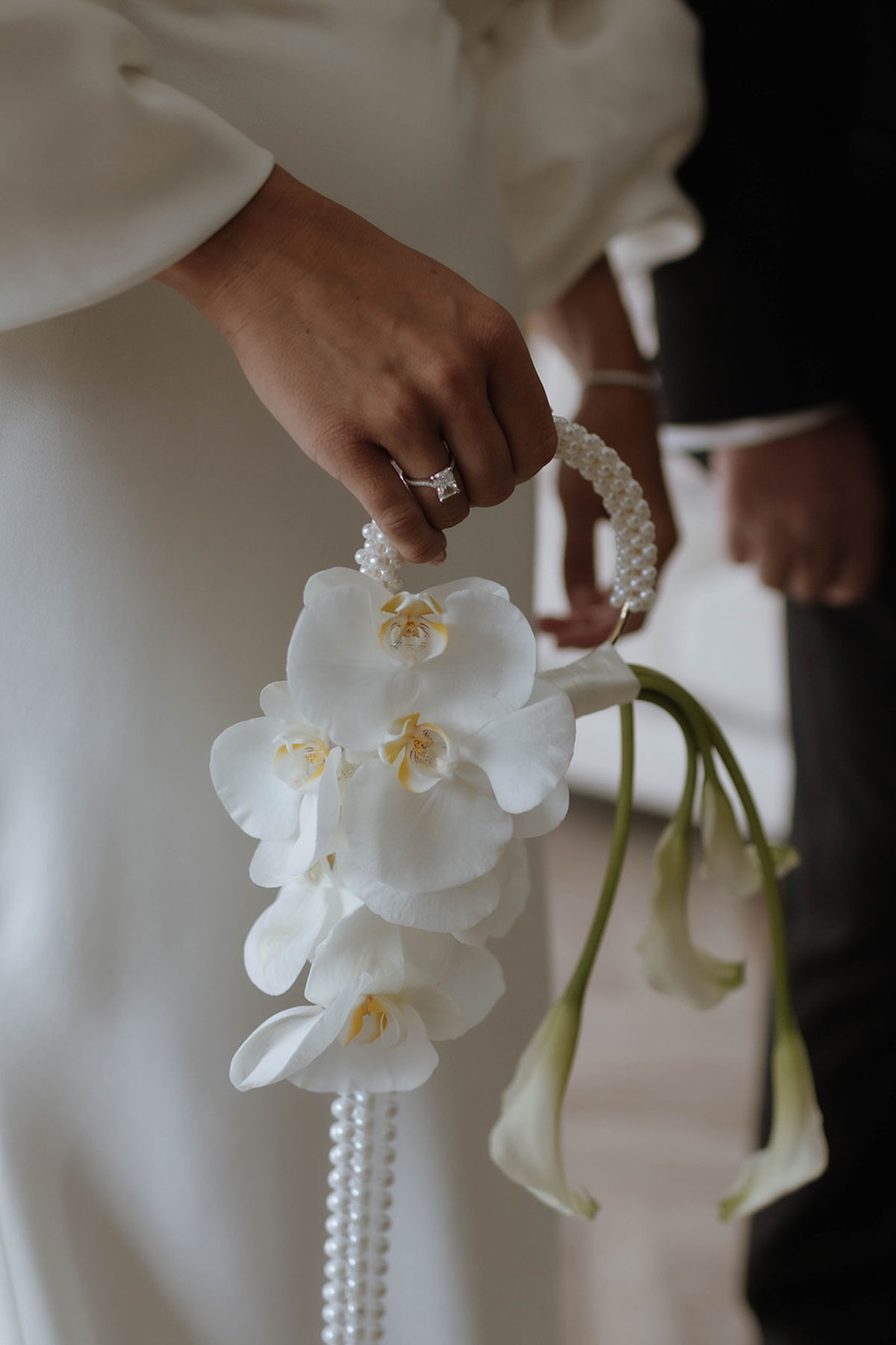 Close-up of bride holding a white orchid bouquet and pearl handle handbag with calla lilies for summer wedding details.