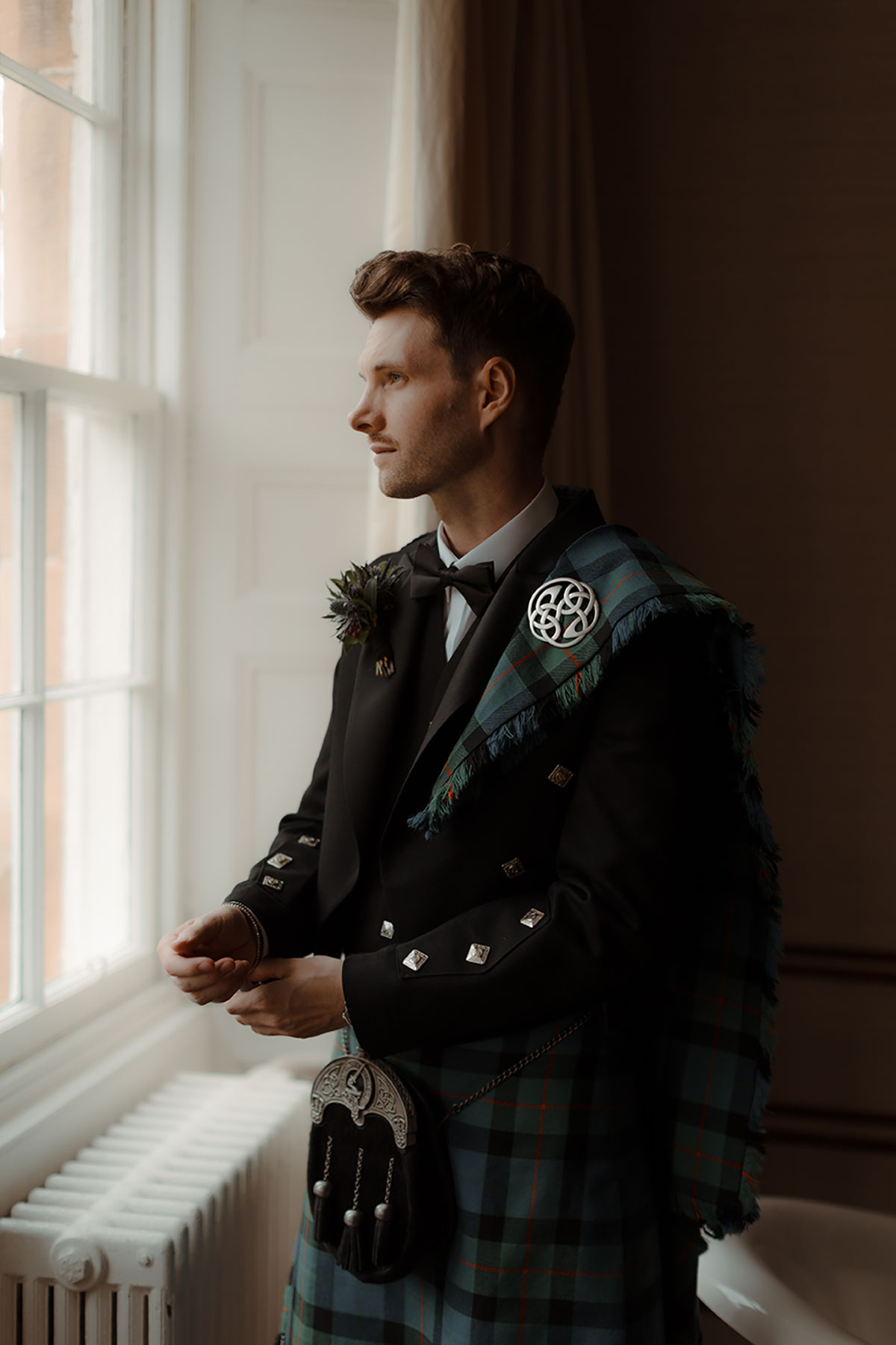 Groom wearing a Gunn tartan kilt with Prince Charlie jacket and fly plaid by MacGregor and MacDuff, standing by a window at Newhall Estate on the morning of his wedding