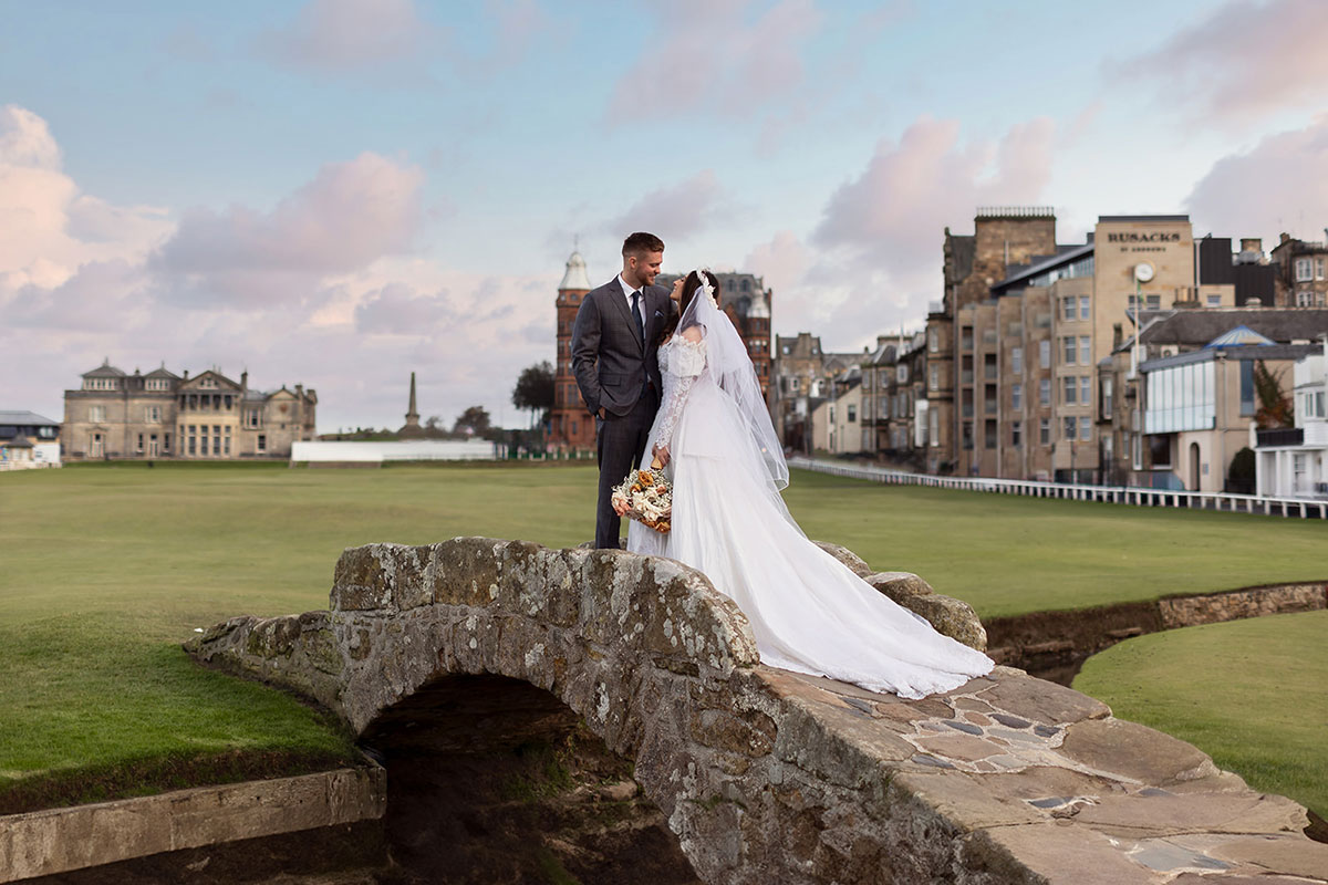 Bride and groom kissing on Swilcan Bridge, St Andrews, with the Old Course and town buildings in the background