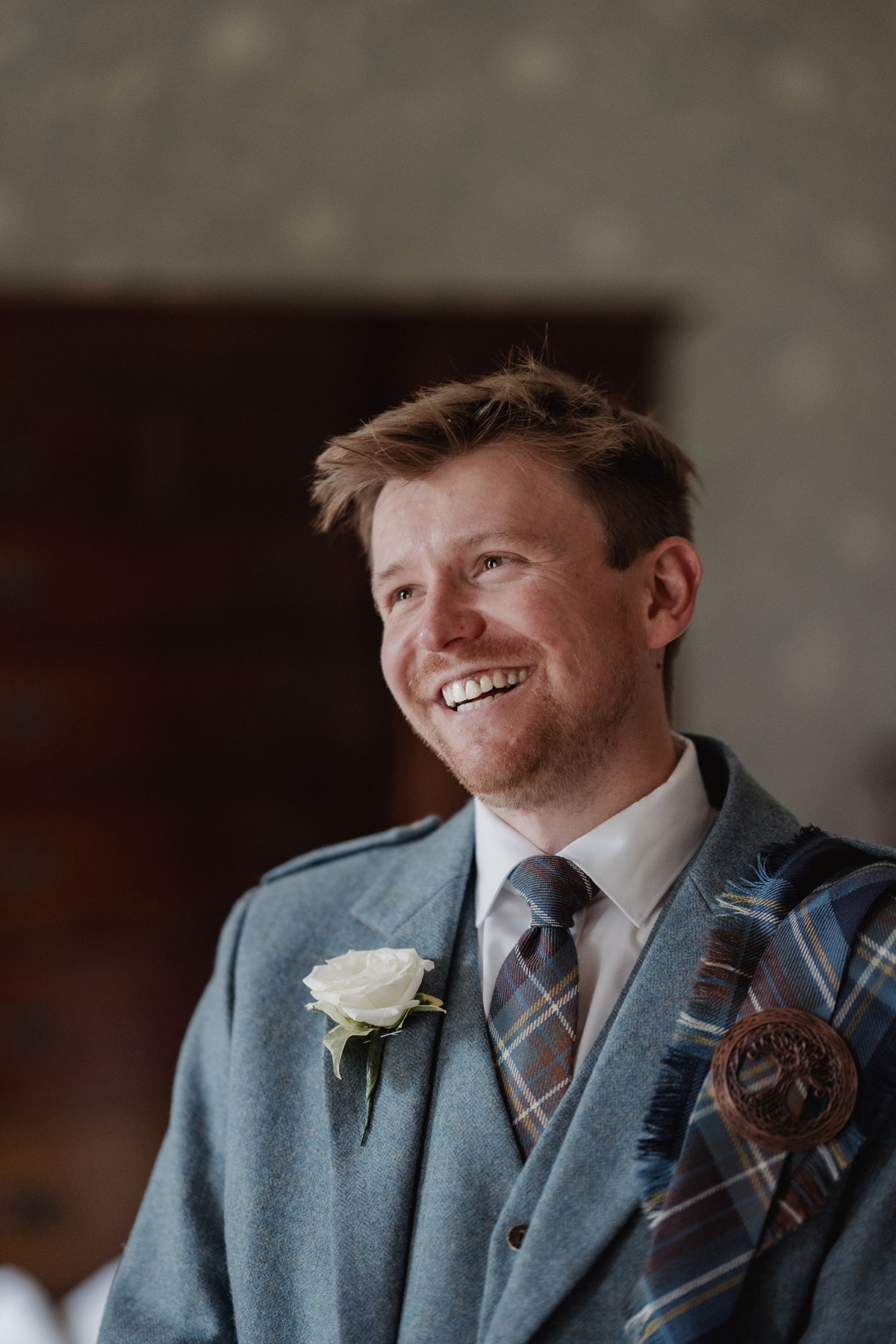 Close-up of a smiling groom wearing a blue tweed kilt jacket, tartan tie, and white rose boutonnière