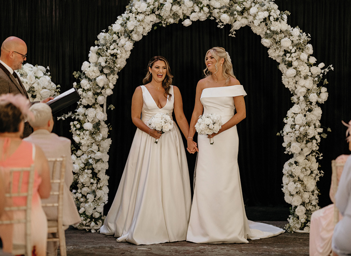 Two brides standing under a white floral arch. They are holding hands and look elated as an officiant in a suit on the left reads from a binder
