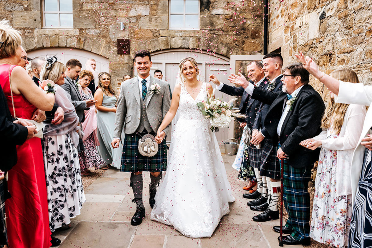 A bride and groom walk hand in hand through a confetti tunnel at Falside Mill, surrounded by cheering guests in colourful outfits and kilts