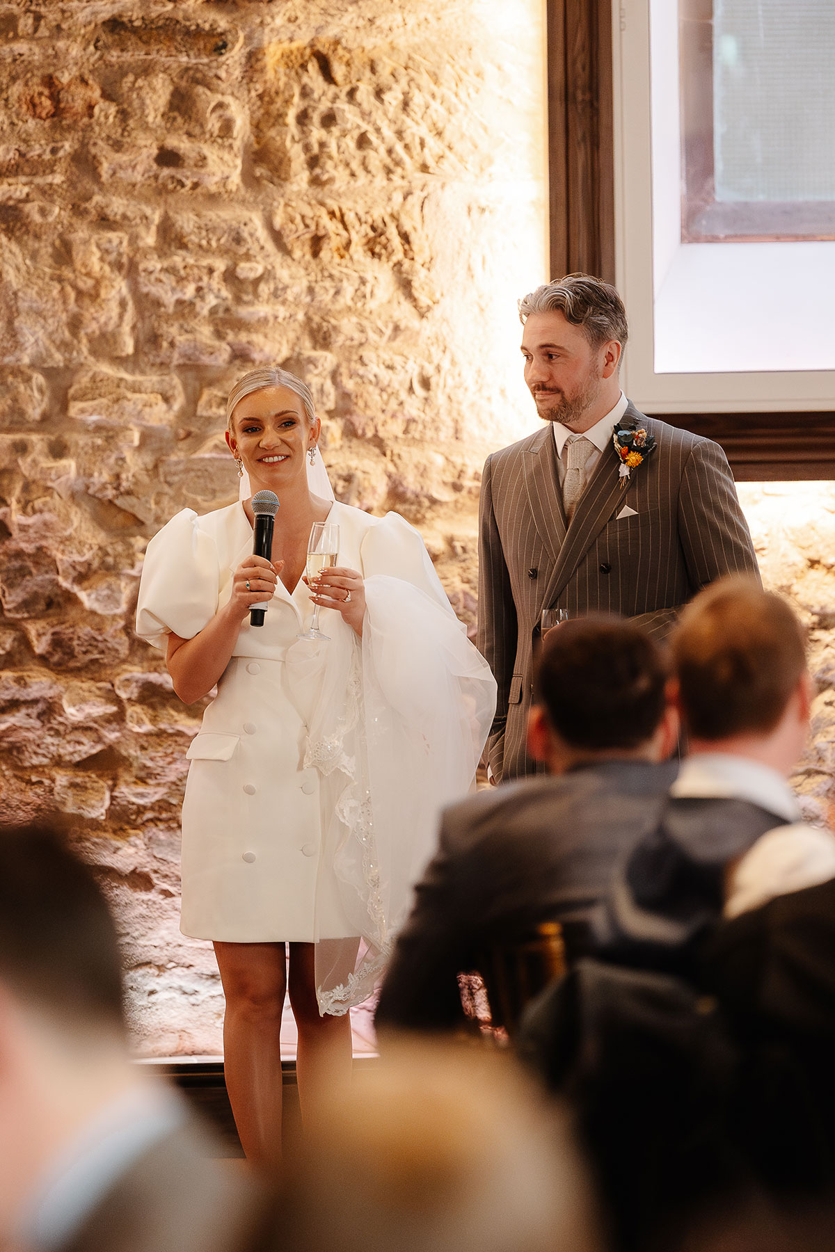 Bride and groom give their wedding speeches at Virgin Hotels Edinburgh’s Greyfriars Hall, standing against the stone wall with a microphone and champagne glass.