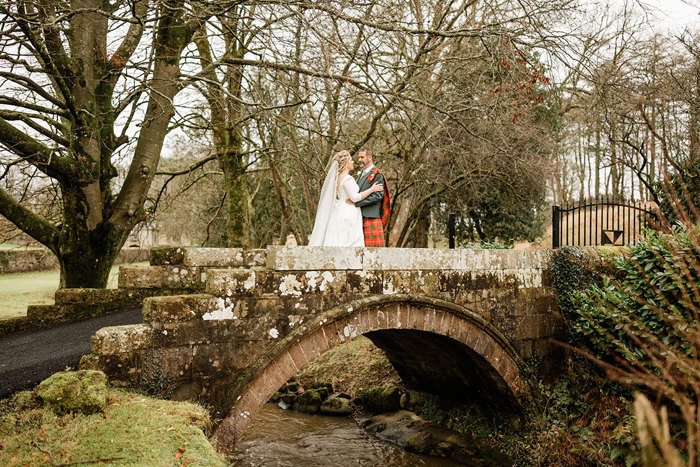 bride in white wedding dress and veil and groom in red and green tartan kilt stand facing each other on brick bridge