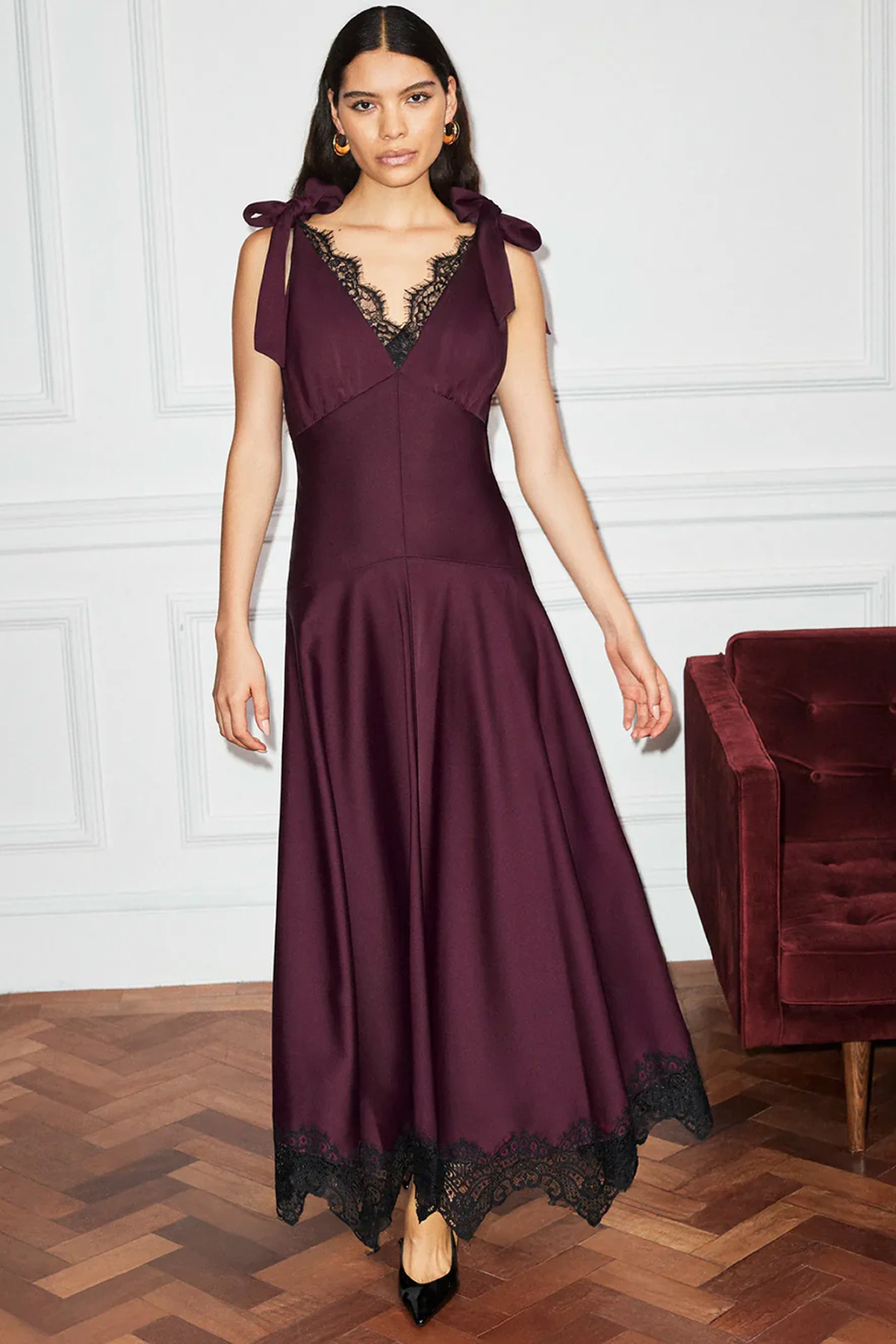 Woman wearing a plum maxi dress with black lace trim, ribbon shoulder ties and a flowing skirt.