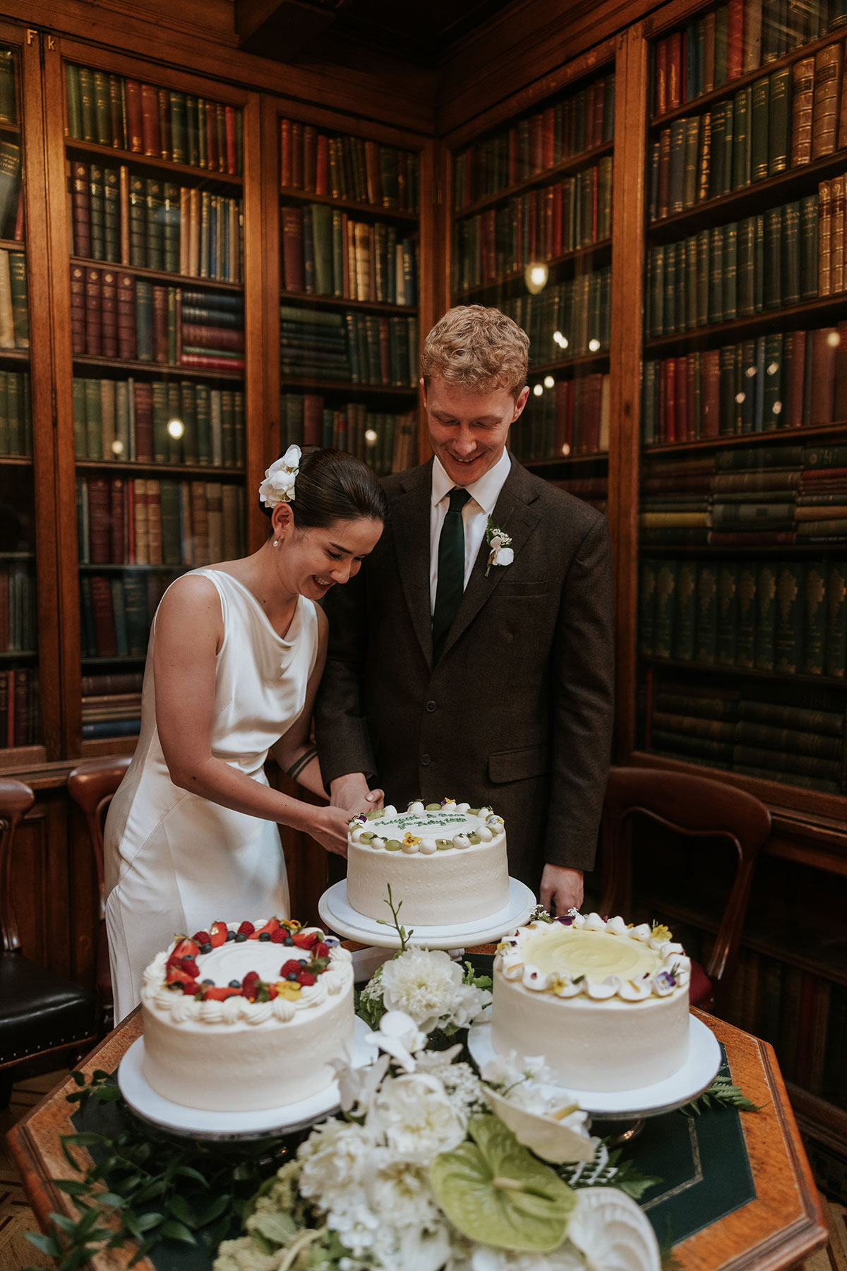 Bride and groom cutting a wedding cake decorated with flowers inside a library venue.