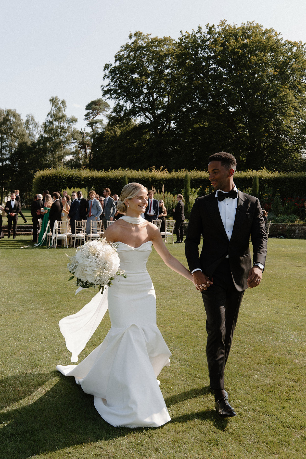 Bride and groom walking hand in hand across the lawn after their outdoor Scottish wedding ceremony, with guests celebrating behind them