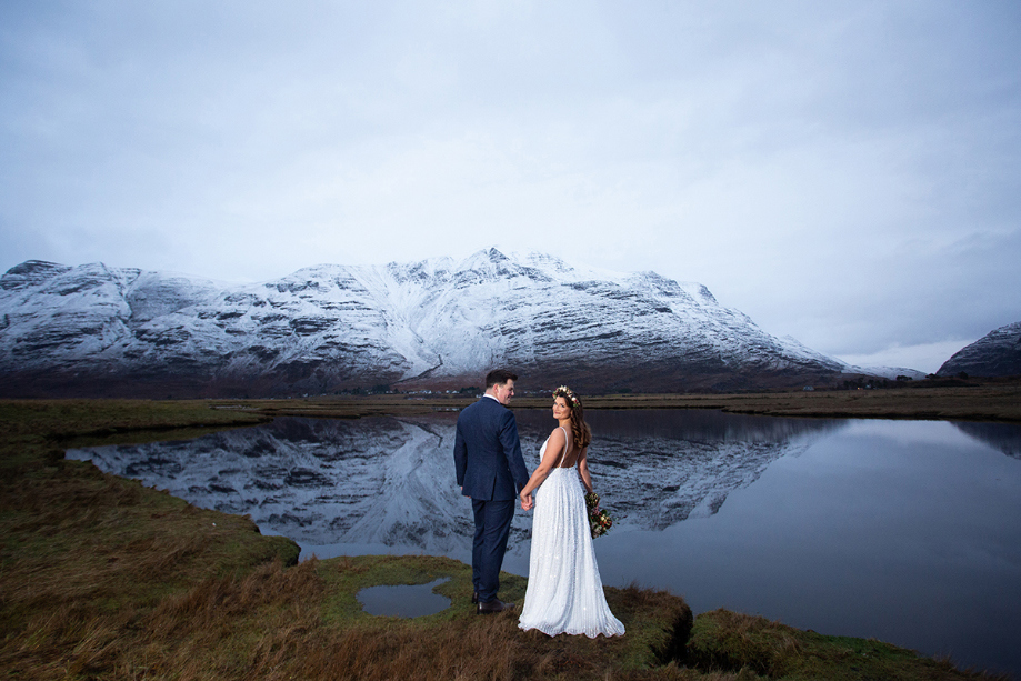 Bride and groom hold hands in front of loch and snow-capped mountain