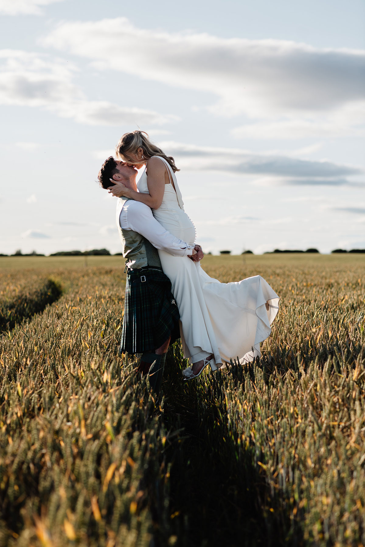 Groom in a kilt lifts the bride in a white dress as they kiss in the middle of a wheat field under a bright sky