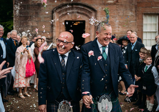 two grooms wearing kilts walk hand in hand while being showered by guests in flower confetti outside Brodick Castle