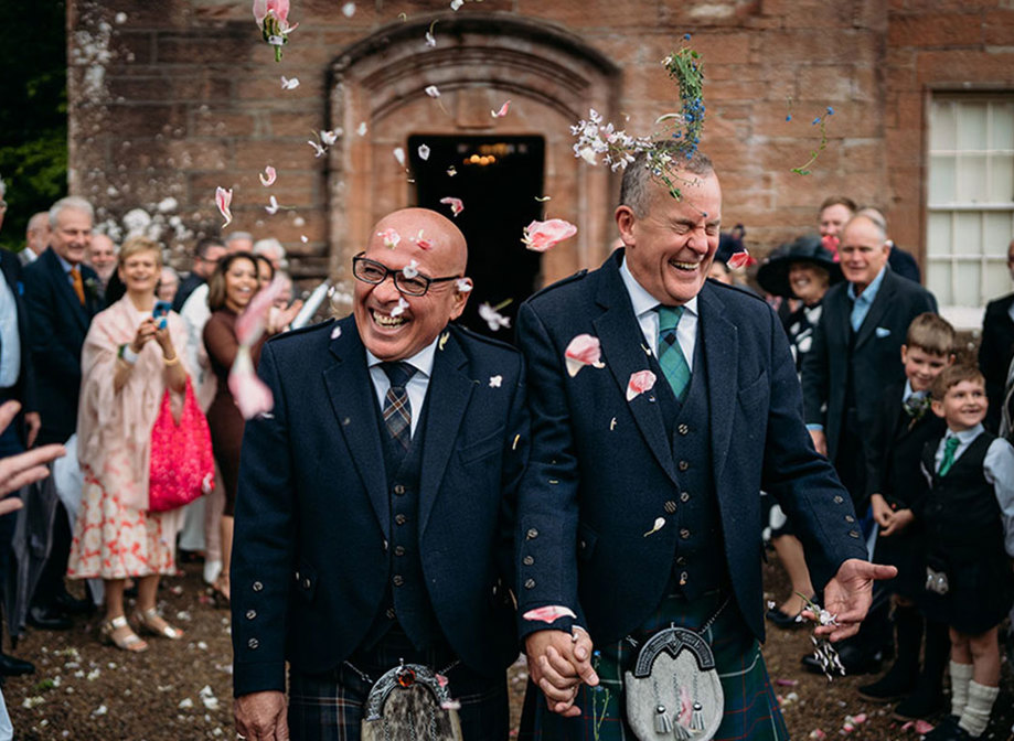 two grooms wearing kilts walk hand in hand while being showered by guests in flower confetti outside Brodick Castle