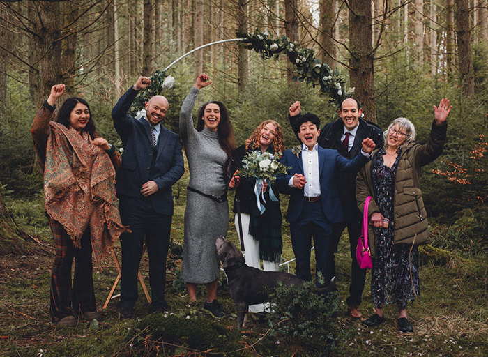 A group of seven people in a forest in front of a floral moongate with their hands up in celebration 
