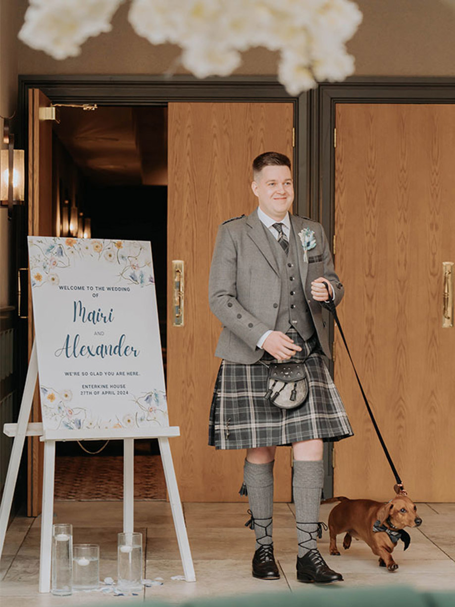 A man in a kilt walks a sausage dog on a lead past a sign that reads 'Welcome to the wedding of Mairi and Alexander'