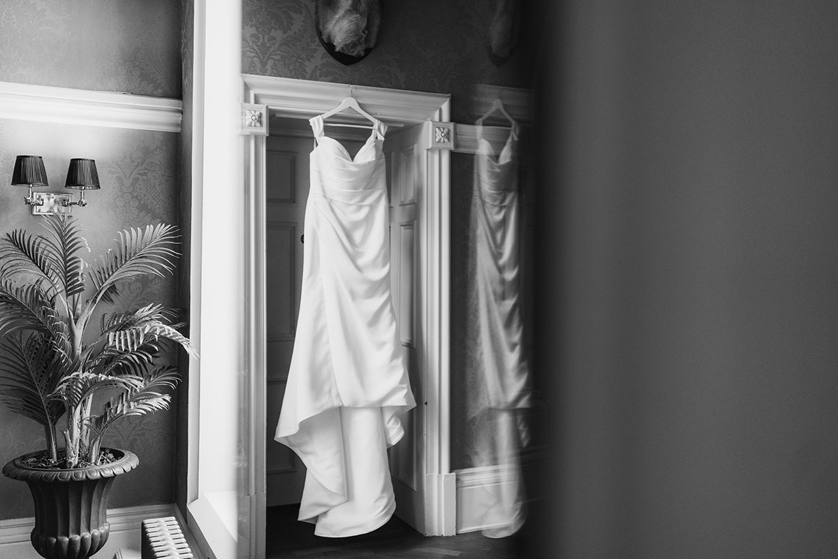 Black and white photograph of a bridal gown hanging in a doorway with its reflection captured in a mirror