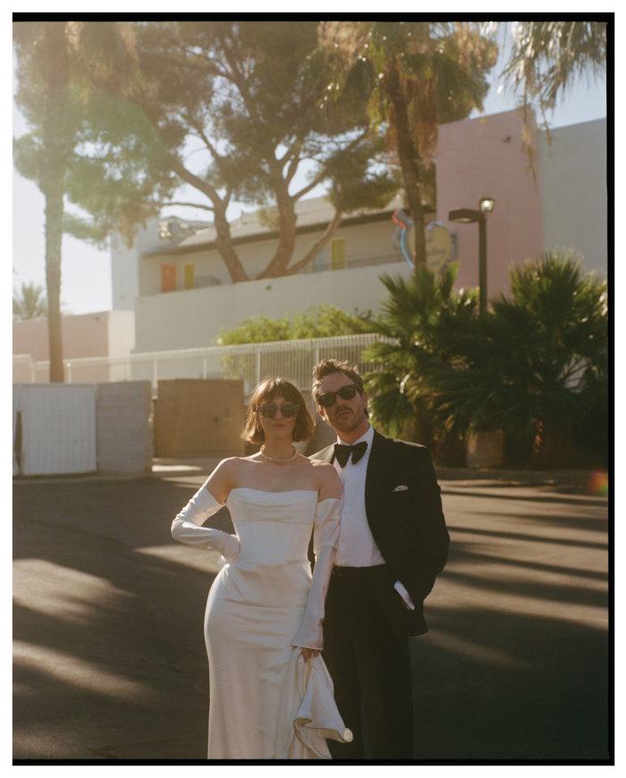 A couple dressed in formal attire stands together outdoors, with palm trees and a building in the background