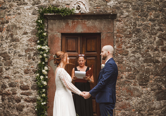 A bride and groom holding hands during a wedding ceremony in front of a rustic brown door and stone wall with an officiant holding a red folio binder in between them