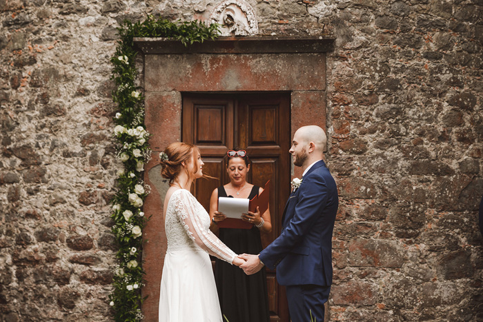 A bride and groom holding hands during a wedding ceremony in front of a rustic brown door and stone wall with an officiant holding a red folio binder in between them