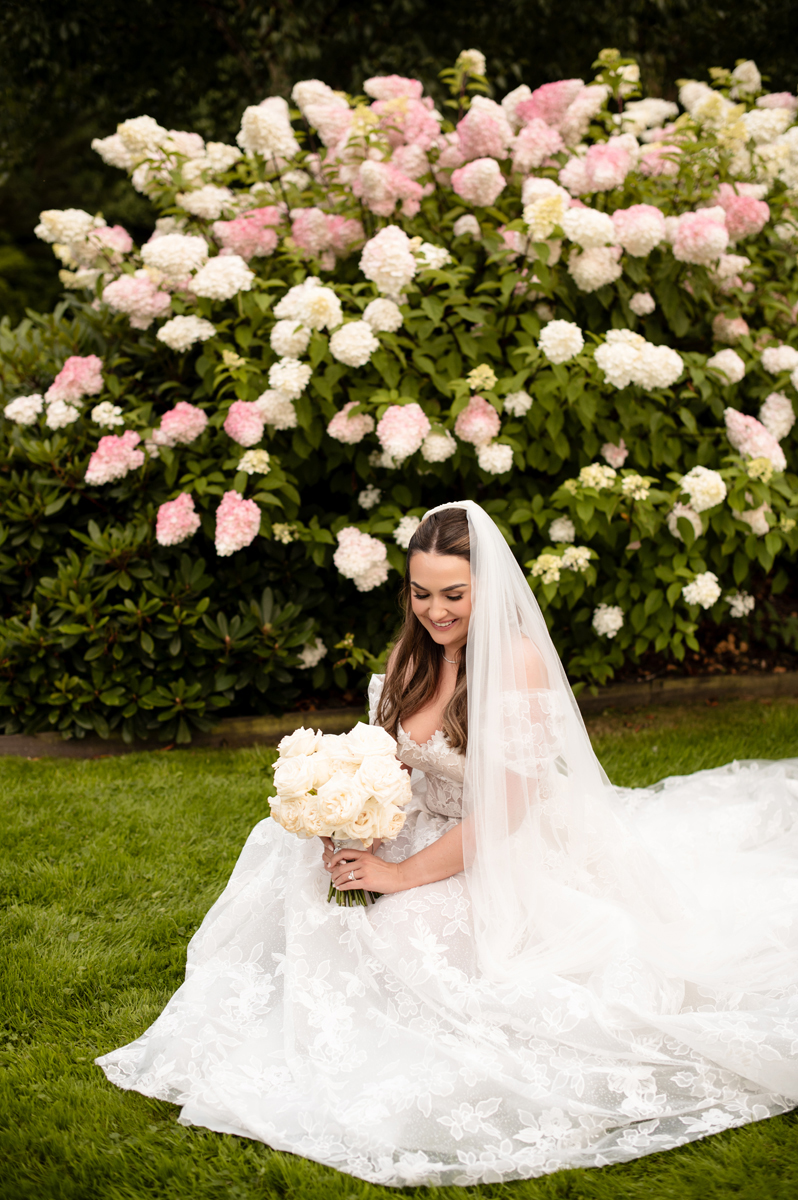 Bride in a floral lace wedding dress and veil holding a white rose bouquet beside hydrangeas in the Crossbasket Castle gardens.