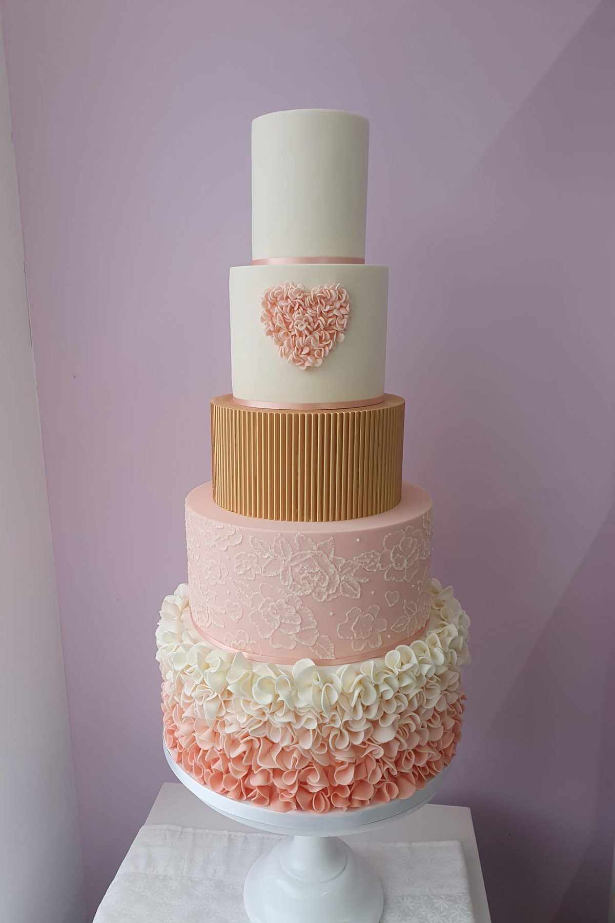 five tier wedding cake in pink, gold and white, with ruffled frosting covering the bottom tier, flowers on the next up, and a pink heart on the second from the top