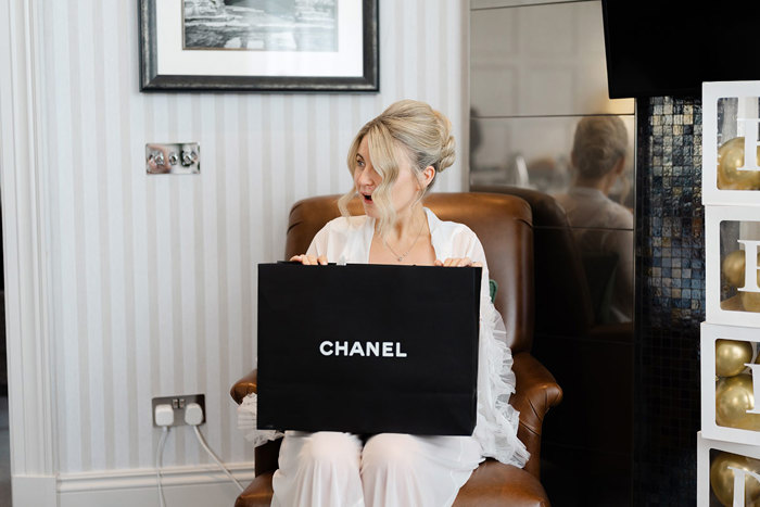 a bride wearing pyjamas seated on a brown armchair with a black Chanel shopping bag on her knee.