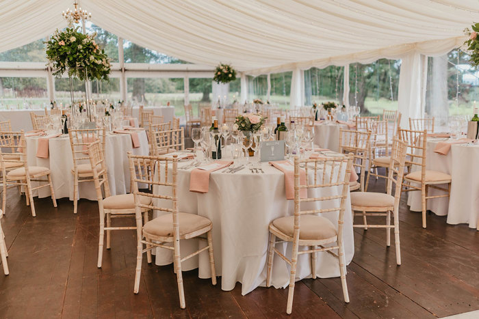The inside of a marquee set up with tables and chairs for a wedding with greenery and pink napkins 