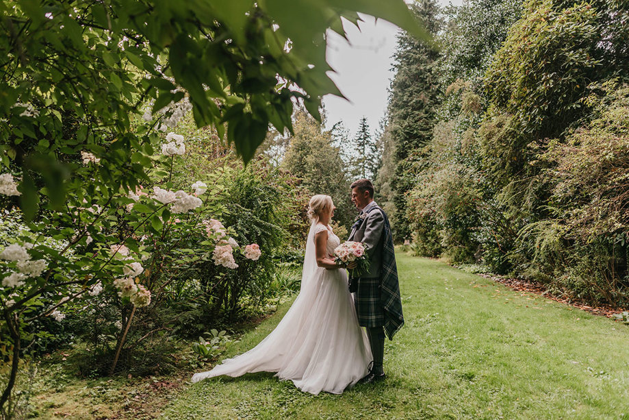 A bride and groom stand in a garden with their arms around each other as the bride holds a white and pink bouquet