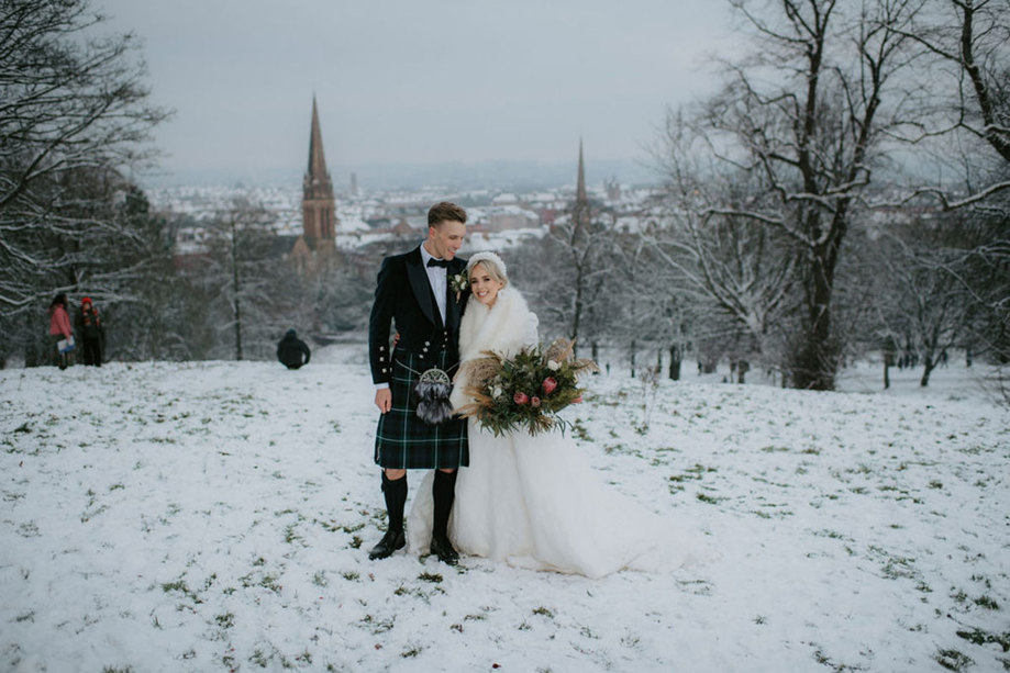 A bride wearing a white dress and white fur wrap standing with a groom wearing a dark kilt in a park in the snow