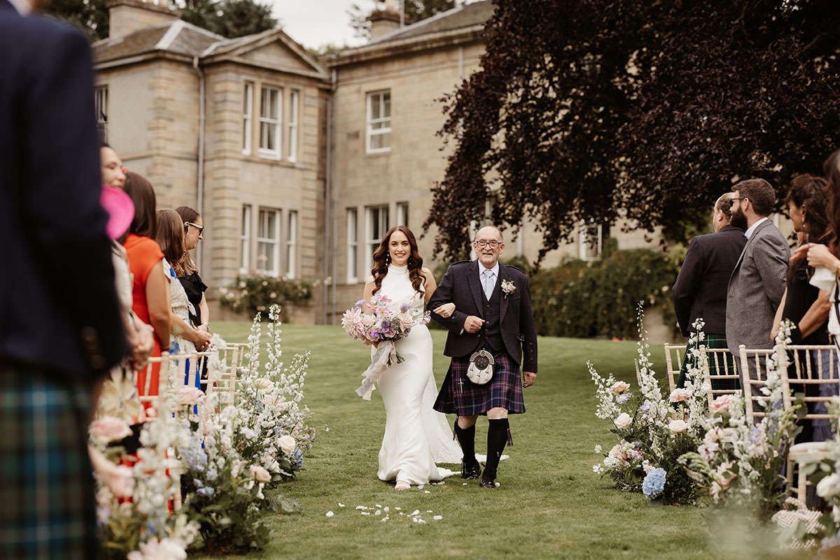 a bride walking down the aisle on arm of a man during an outdoor wedding ceremony at Netherdale House