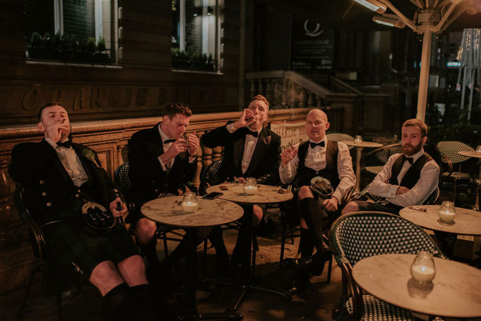 A group of five men wearing kilts sit outside smoking cigars 