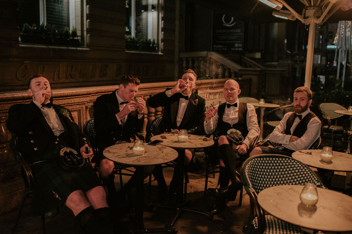 A group of five men wearing kilts sit outside smoking cigars